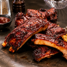 Sticky glazed barbecue ribs stacked on a platter, with sauce dishes in the background