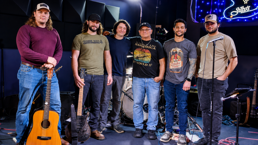 Six musicians stand on a small stage with instruments under blue-purple lights.