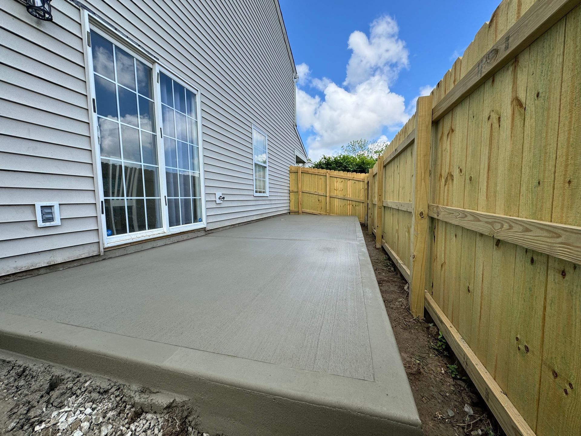 Concrete patio beside a house, bordered by a wooden fence and sliding glass doors under a partly cloudy sky