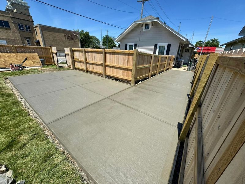 Concrete driveway beside a wooden fence and small house under a clear blue sky