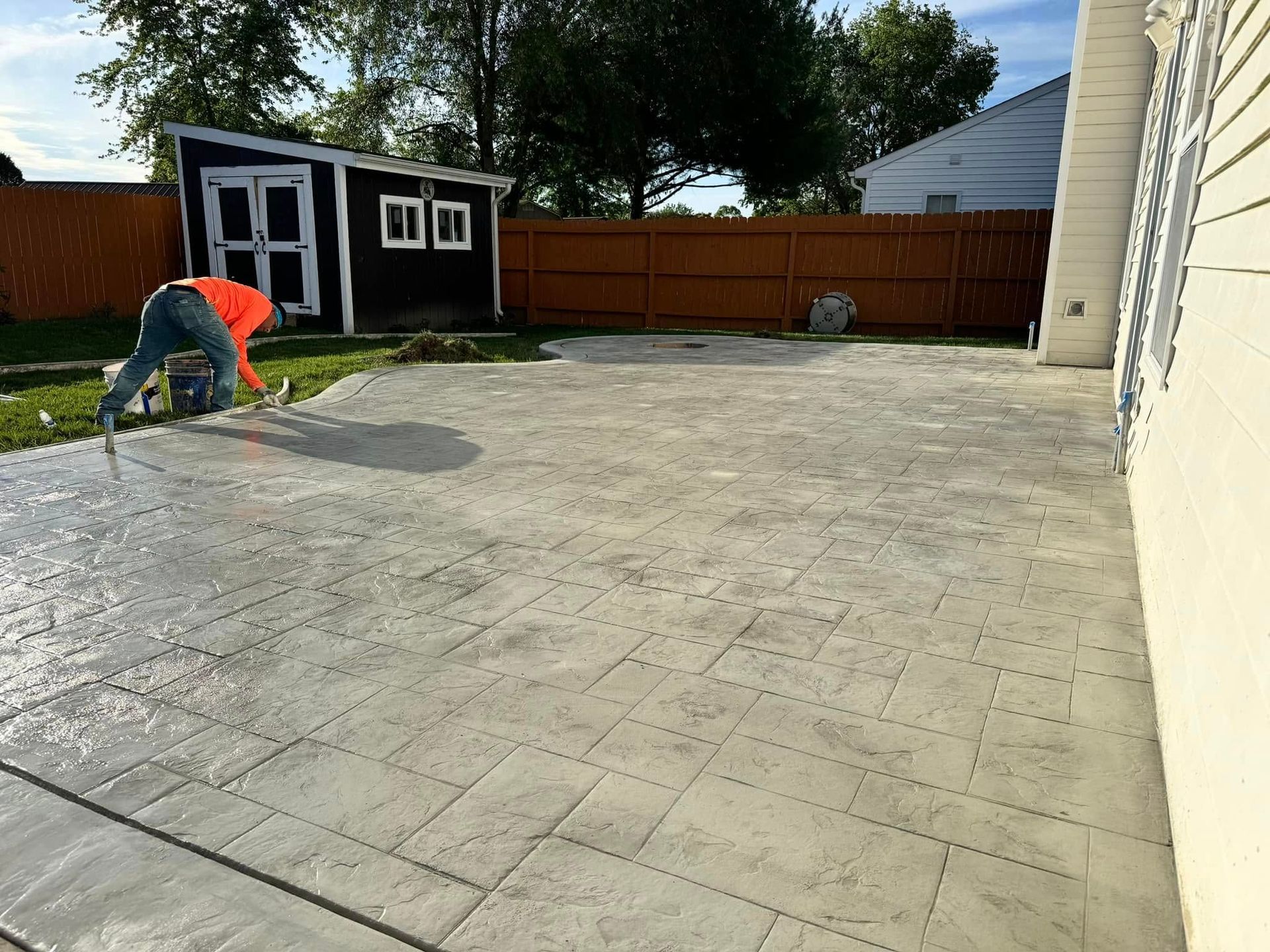 Worker pressure washing a large concrete driveway beside a house and shed in a backyard
