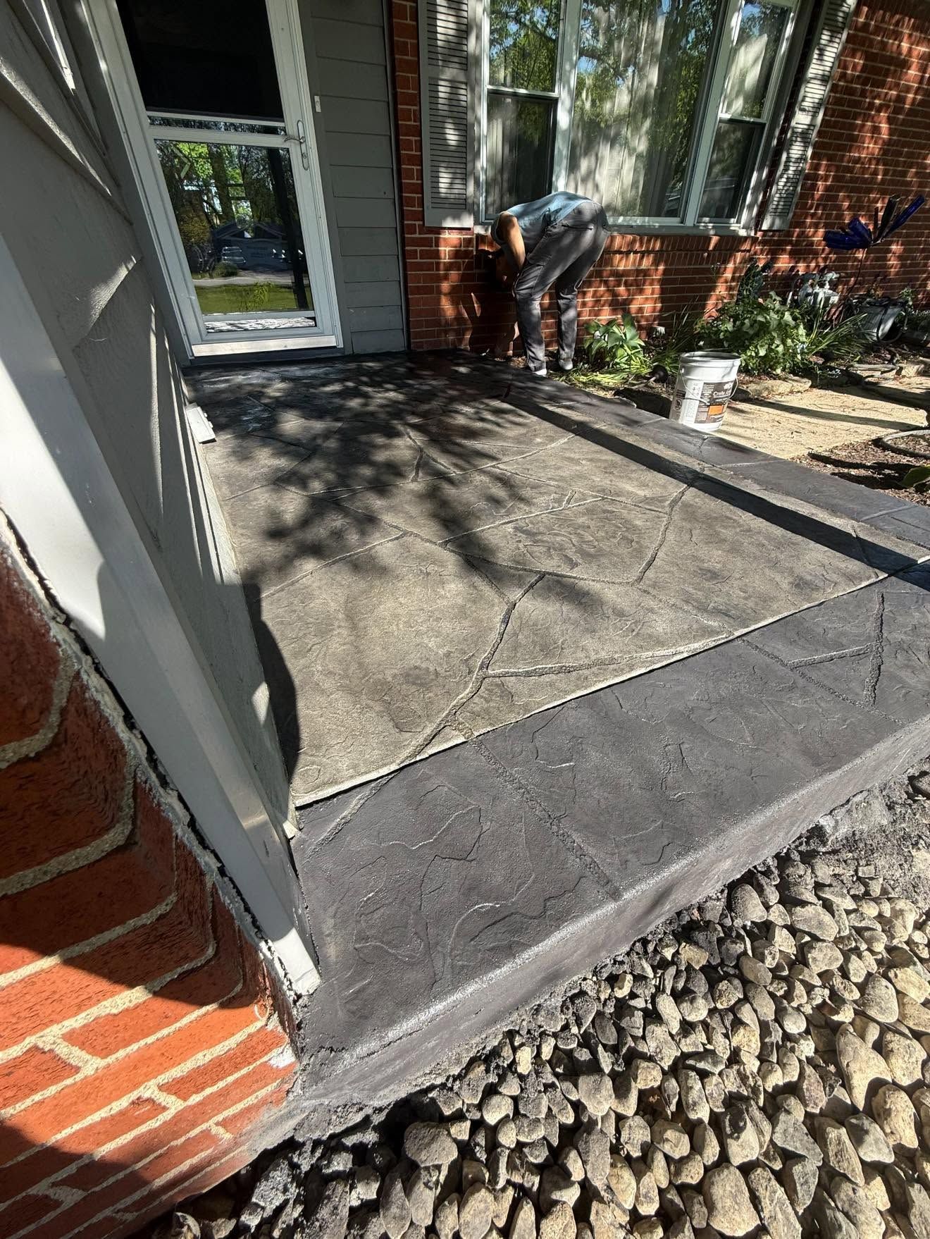 Concrete front porch with a step, glass door, brick wall, and decorative rocks in sunlight