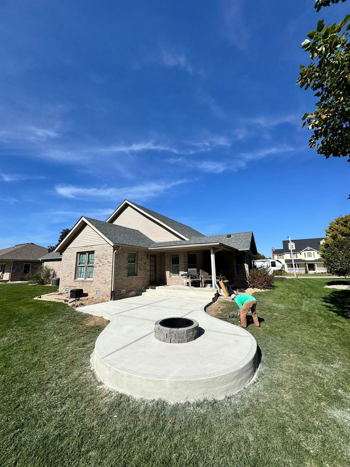 Backyard with circular fire pit patio beside a brick house, under a bright blue sky; person kneels nearby.