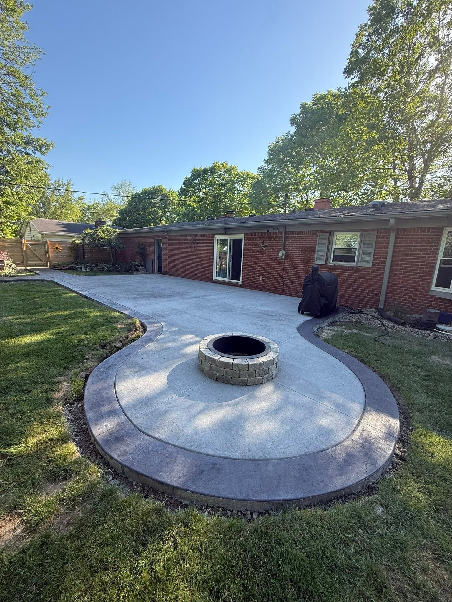Backyard patio with circular stone fire pit, curved paving, brick house, and trees under a blue sky