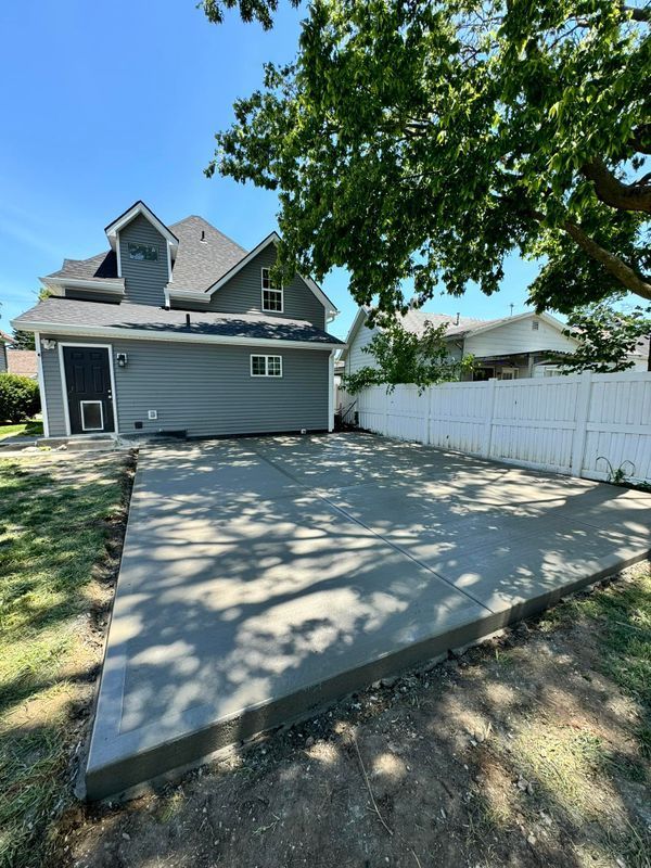 Concrete driveway beside a gray house with a white fence and tree shade