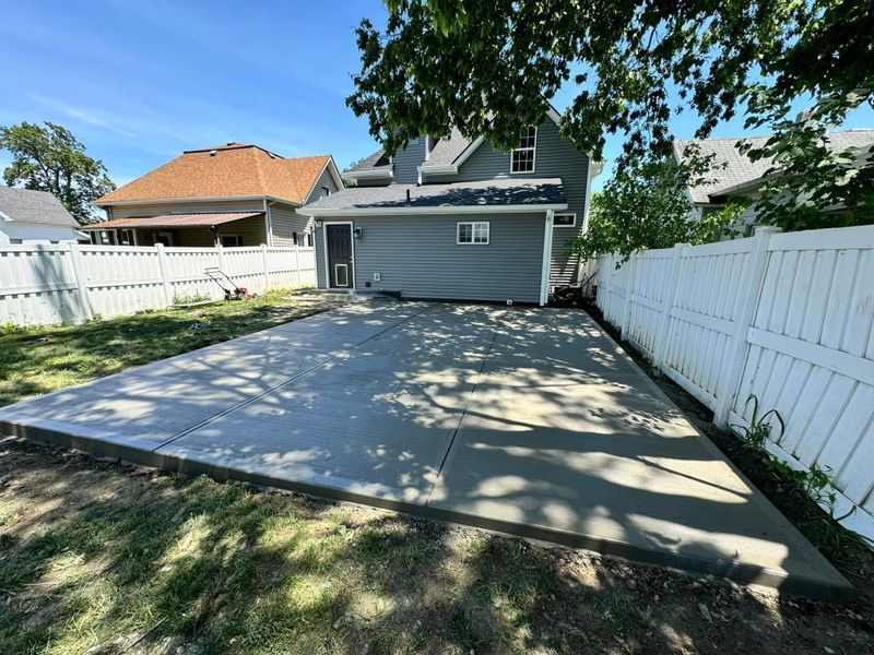 Backyard concrete patio behind a gray house, bordered by white vinyl fences and trees