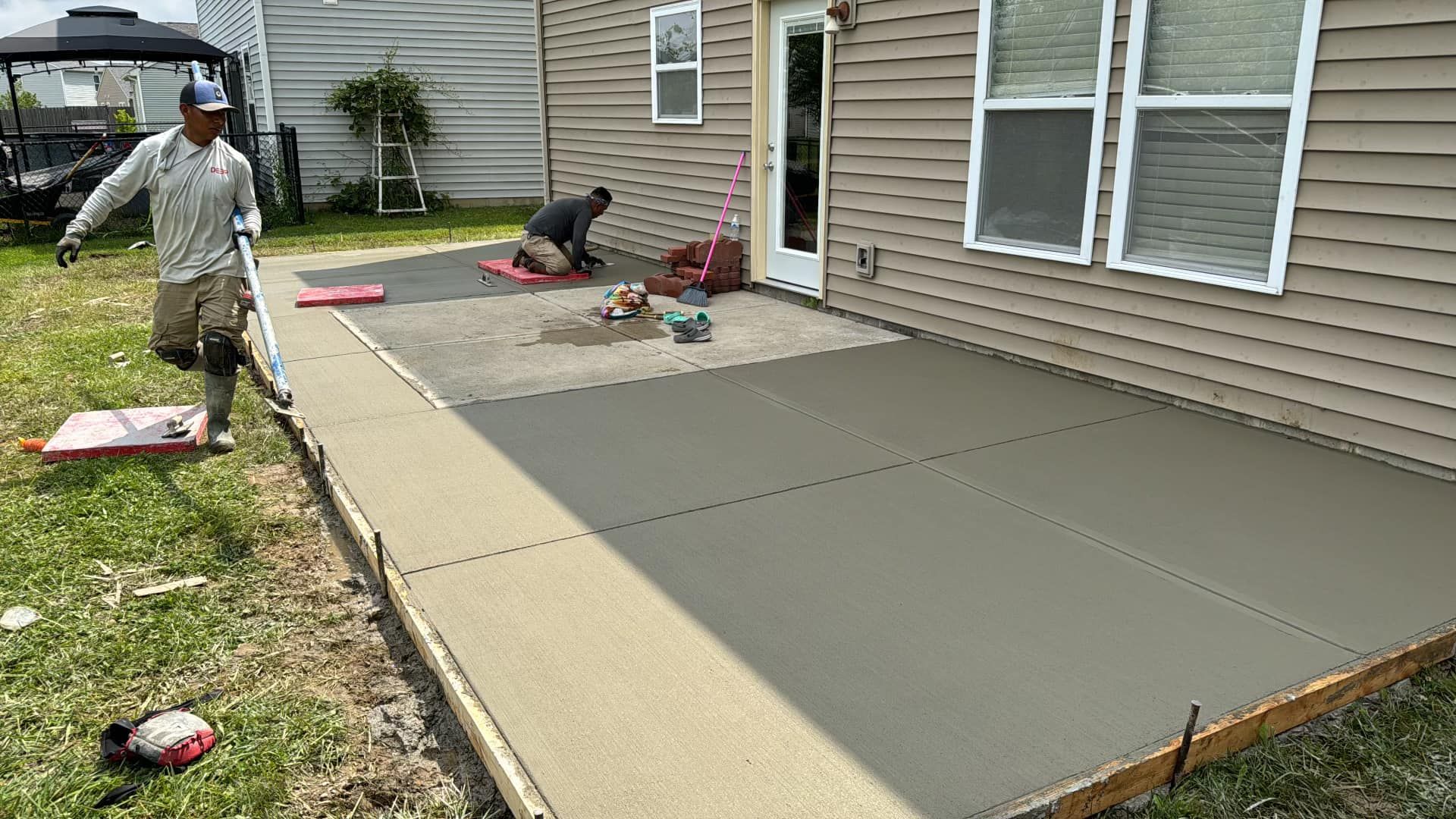 Two workers pour and smooth a new concrete patio beside a house.