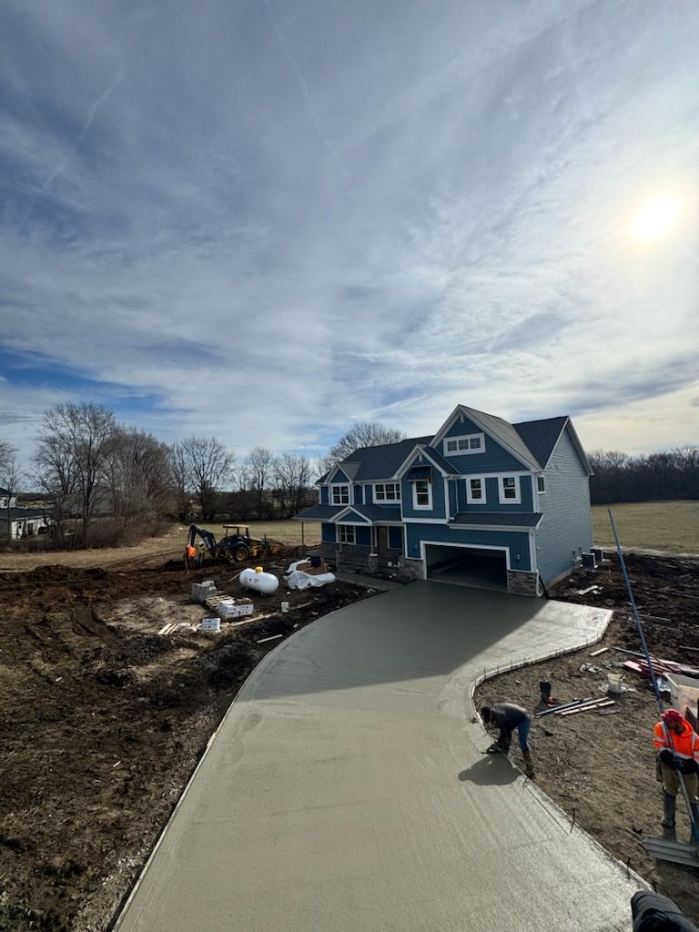 Blue house with unfinished concrete driveway; two workers pour or smooth pavement under a bright sky.
