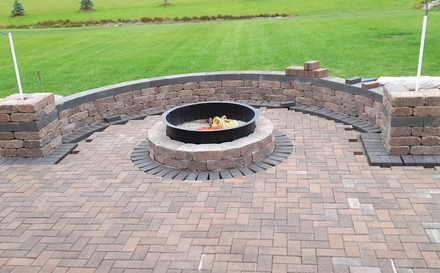Stone patio with a circular fire pit surrounded by low brick seating and a grassy yard beyond