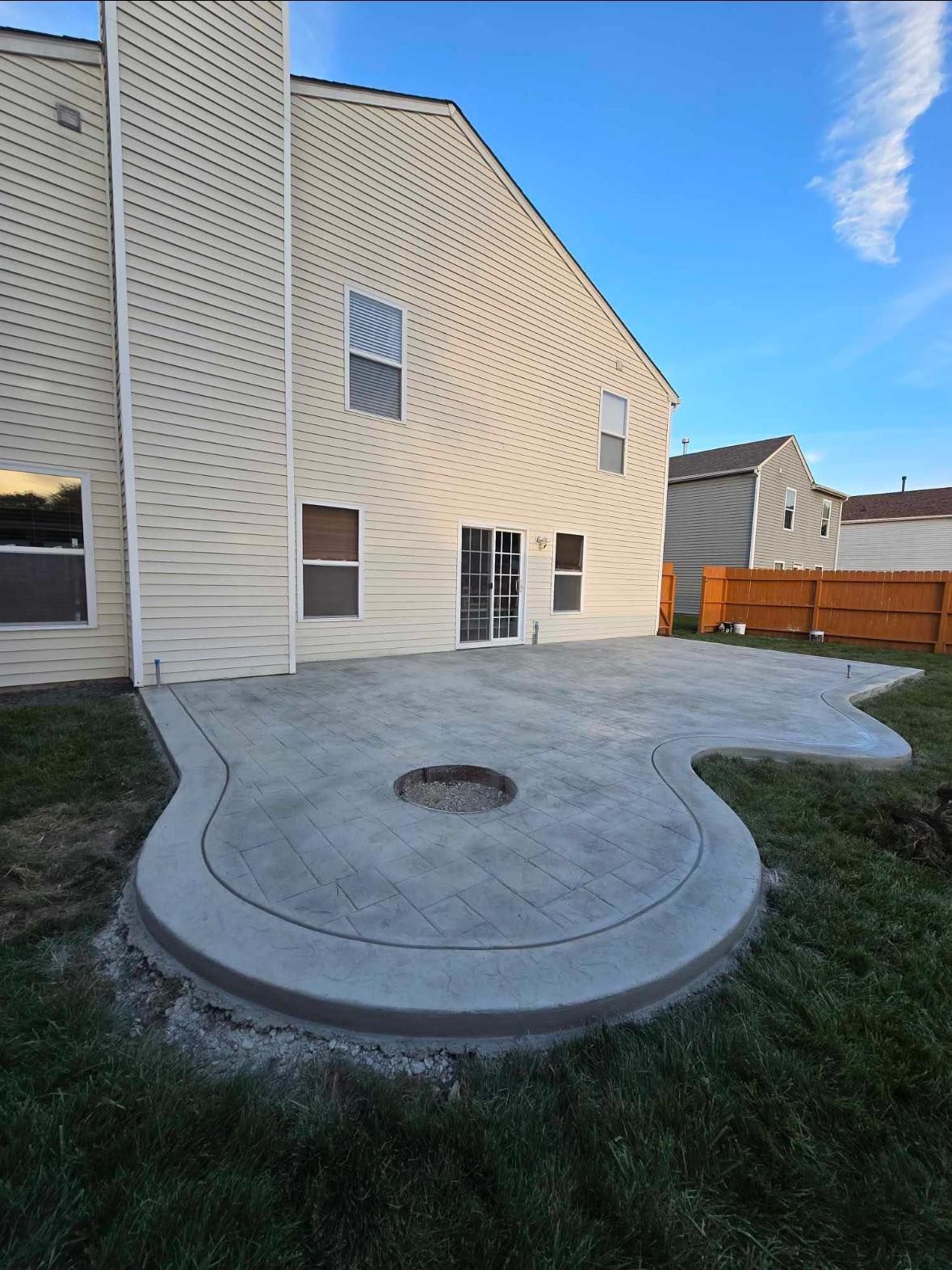Backyard concrete patio with a circular fire pit area beside a beige house and wooden fence