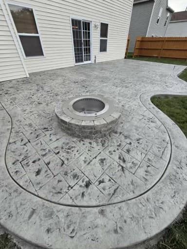 Backyard concrete patio with a circular stone fire pit beside a house and wooden fence.