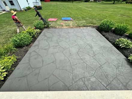 Stamped concrete patio bordered by mulch and plants, with two people working in the lawn nearby.
