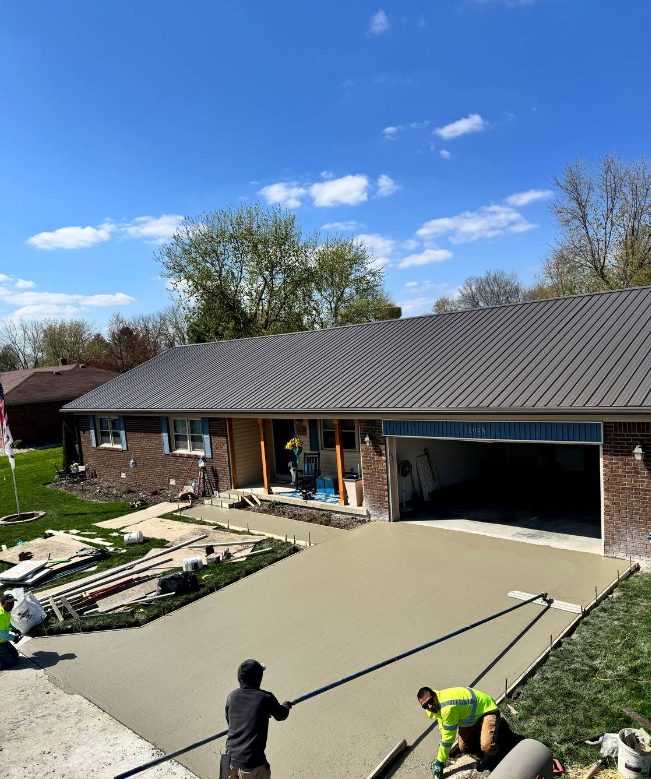 Workers smoothing a fresh concrete driveway beside a brick house and open garage on a sunny day