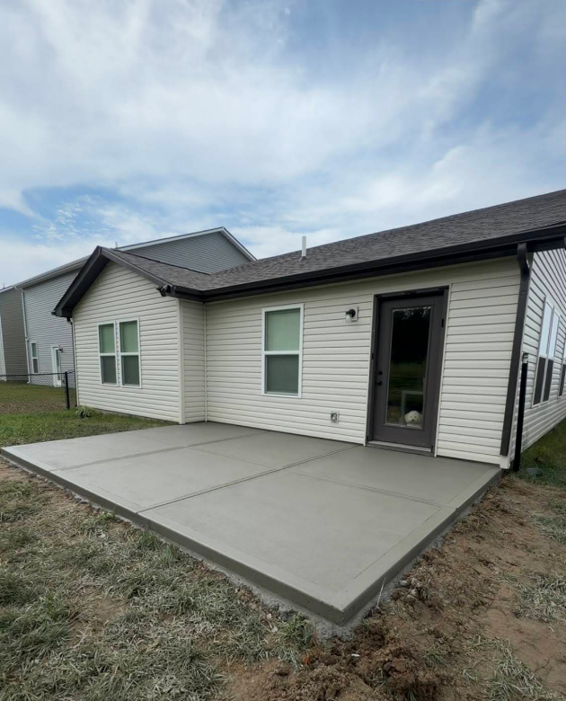 Single-story house exterior with a large concrete patio and grassy yard under a cloudy sky