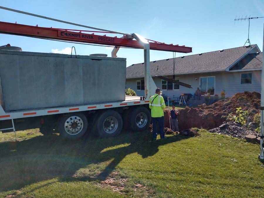 A concrete septic tank is lifted by a crane on a trailer next to a house with a hole in the yard.