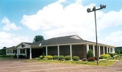Single-story brick building with covered porch, shrubs, and parking lot under a cloudy sky