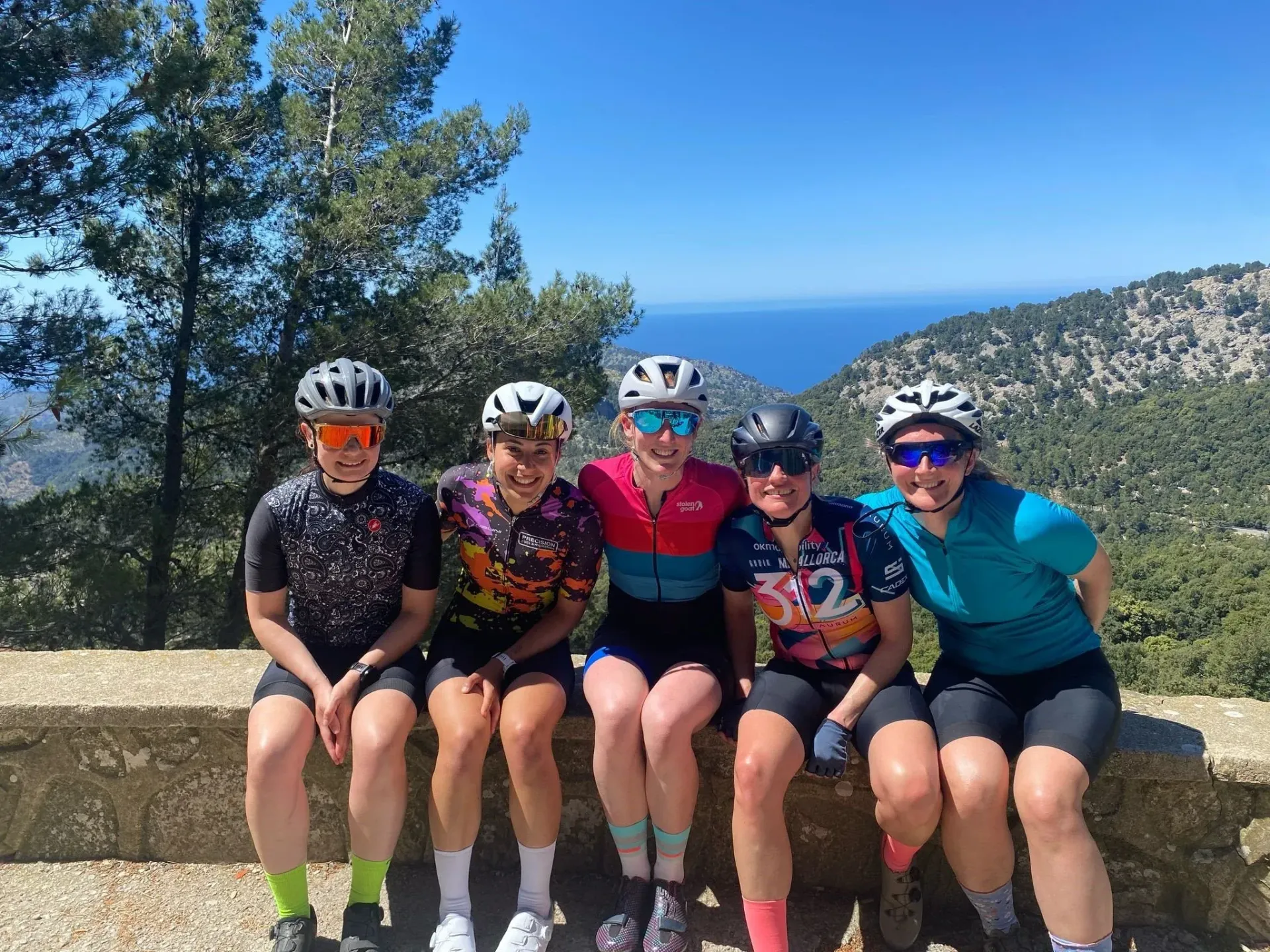 Five cyclists in colorful gear sit on a stone wall with mountain and lake views behind them.