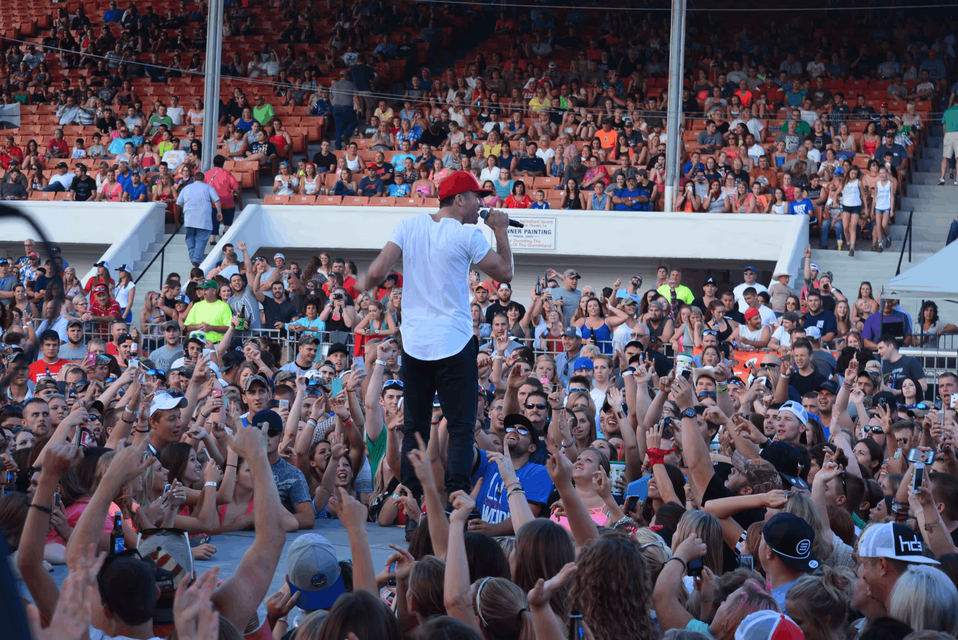 Singer in a red cap performs onstage before a cheering crowd with raised hands in a packed stadium