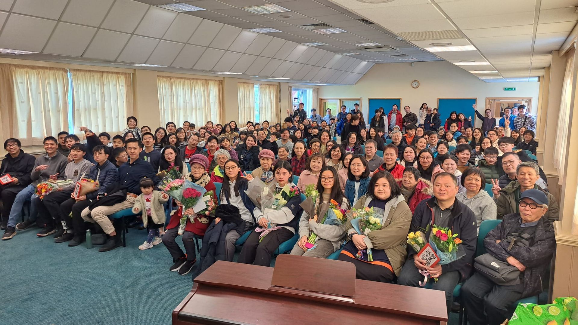 A large group of people are sitting in a room holding flowers.