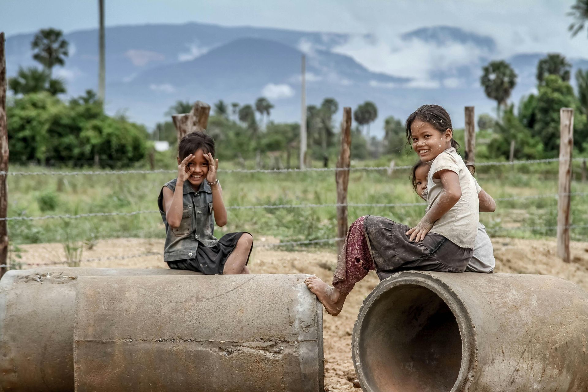 Three children are sitting on concrete pipes in a field.