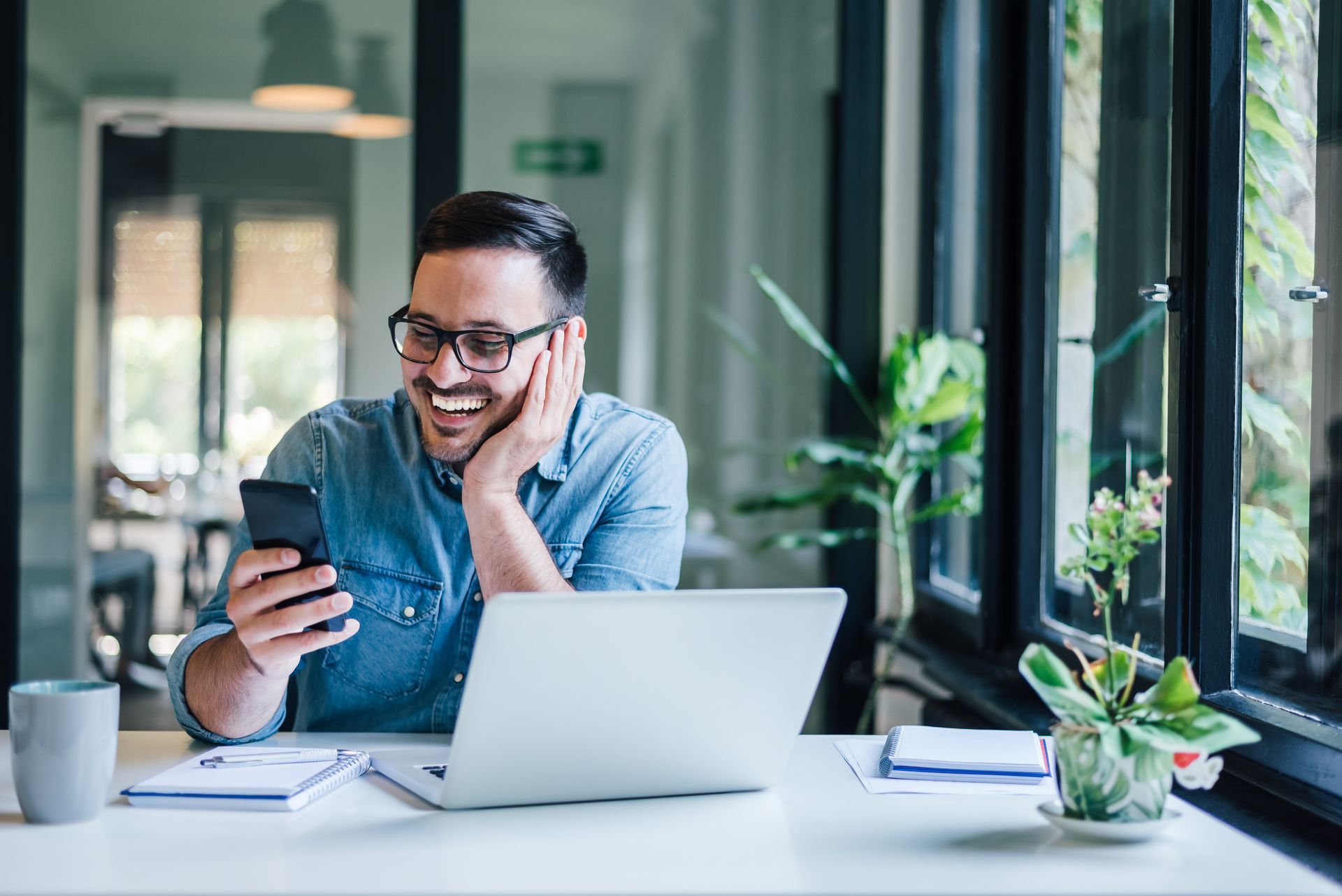 Man with glasses smiling while looking at his phone, sitting at a desk with a laptop, window, and plants.