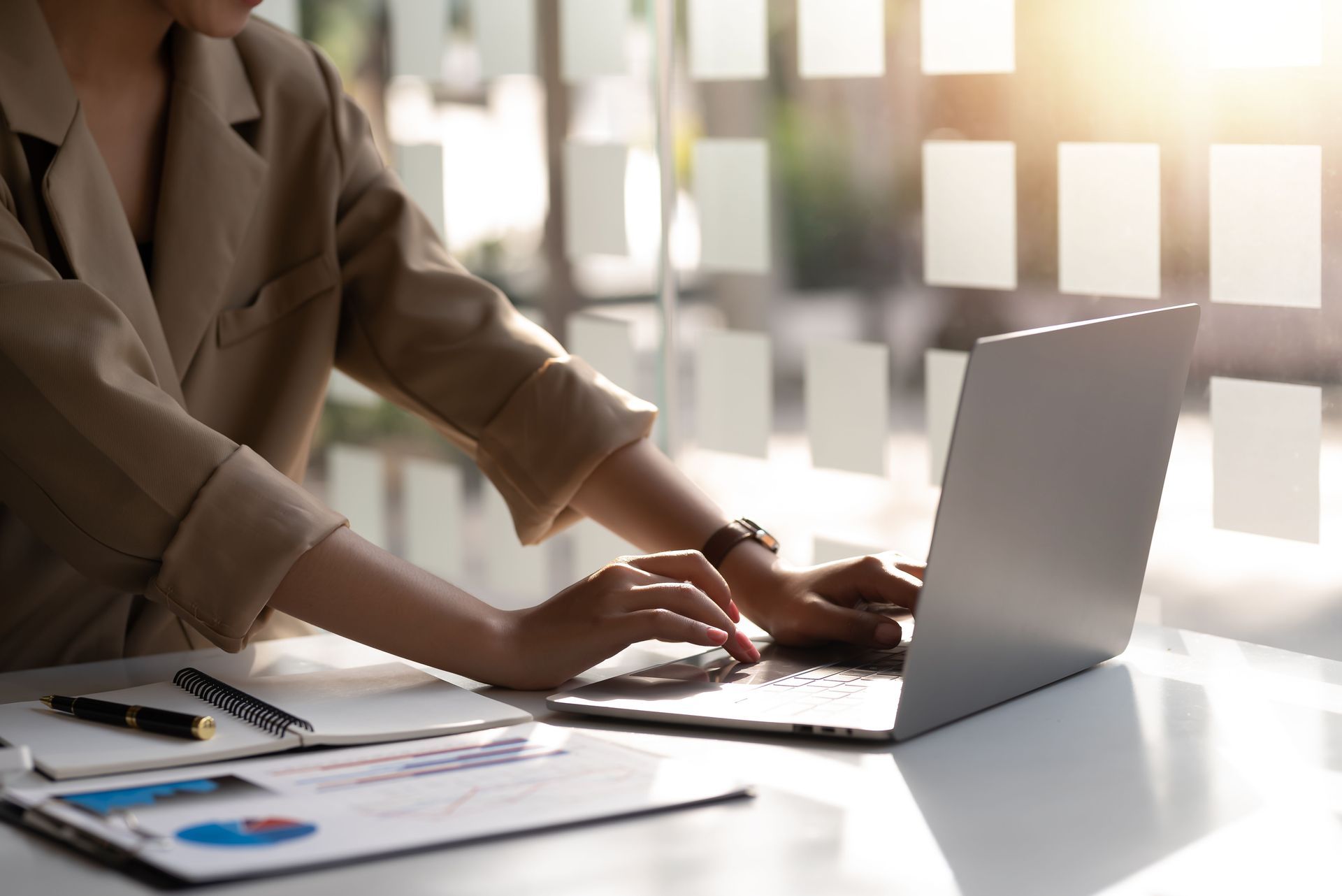 A person in a tan blazer is typing on a laptop next to documents and a notebook on a bright office desk.