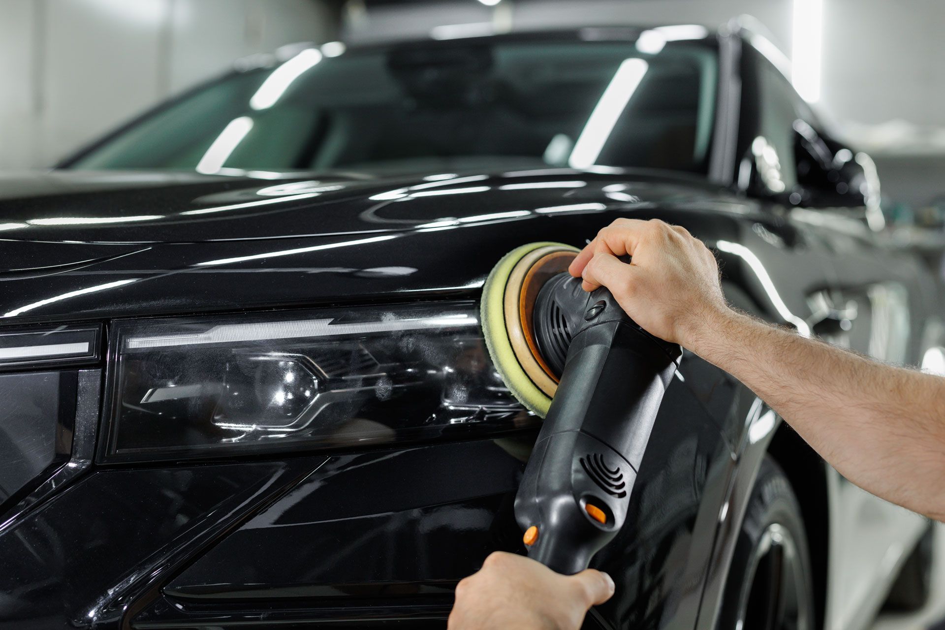 Person polishing a black car with a rotary buffer in a well-lit shop.