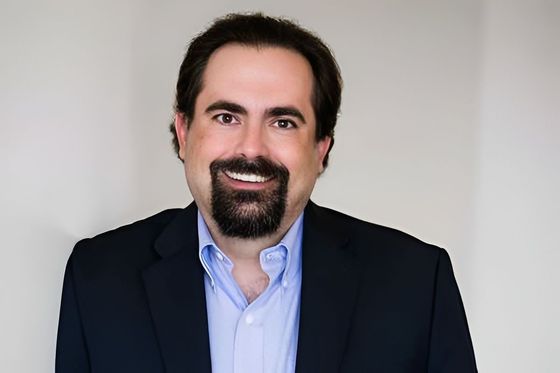 Man with dark hair and beard, wearing a blue shirt and navy blazer, smiles at the camera.