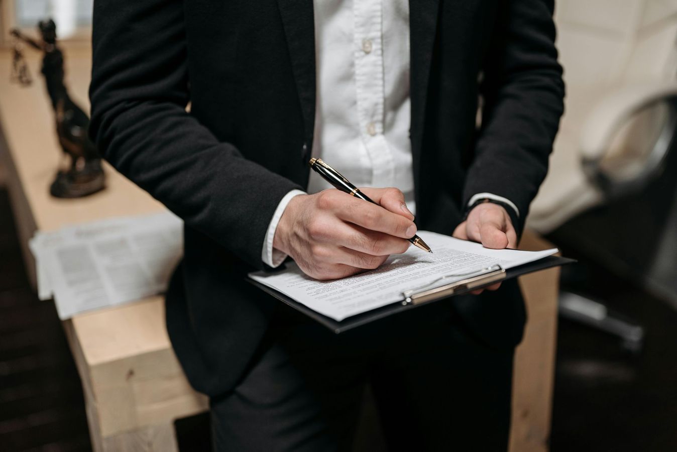 Man in suit writing on a clipboard, leaning on a desk in an office.