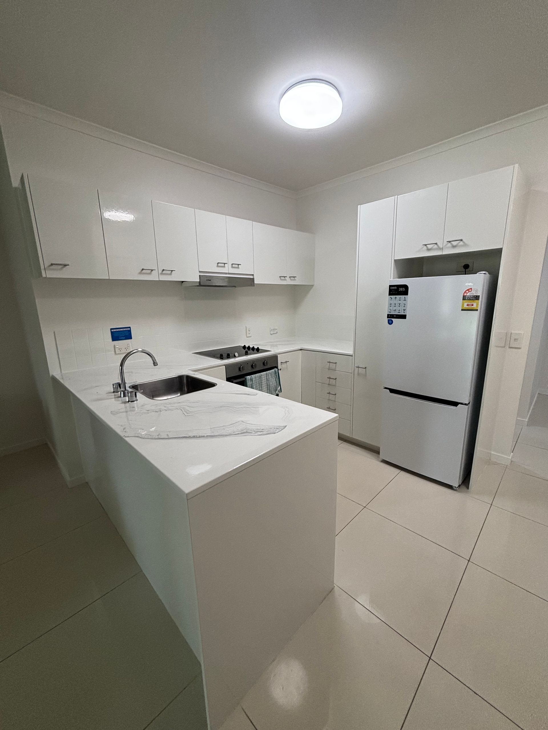 Bright white compact kitchen with an island, cabinets, sink, and refrigerator under a ceiling light. — Supremacy Painting Co In White Rock, QLD