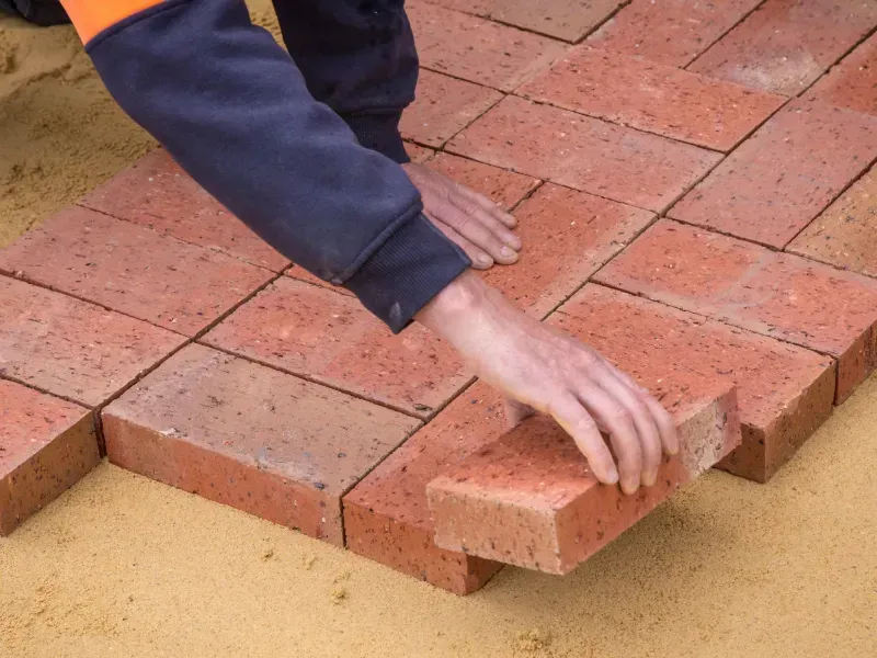 Person laying red bricks in a herringbone pattern on a bed of sand.