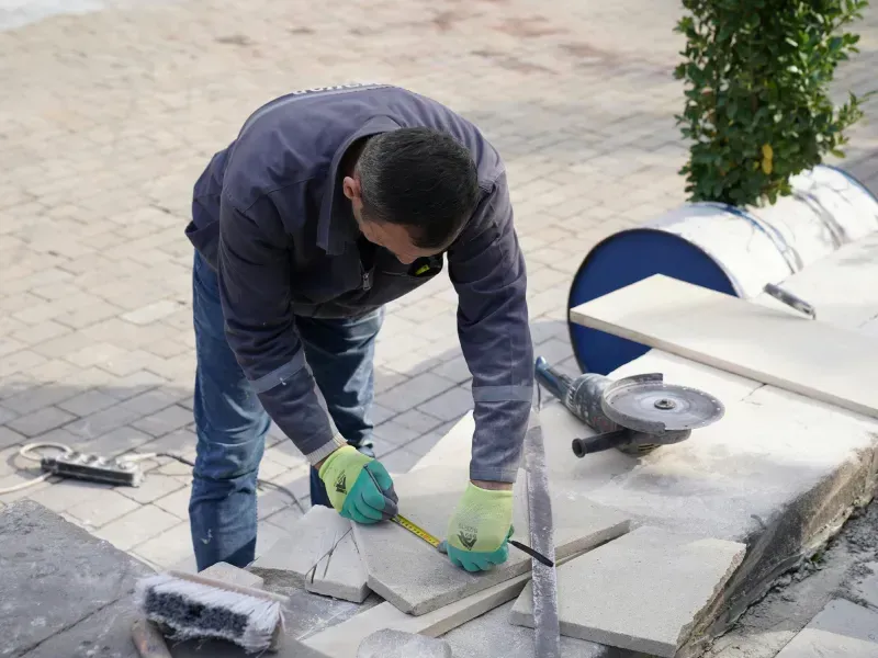 Person using a tape measure to measure a stone tile, with a grinder nearby, outdoors.