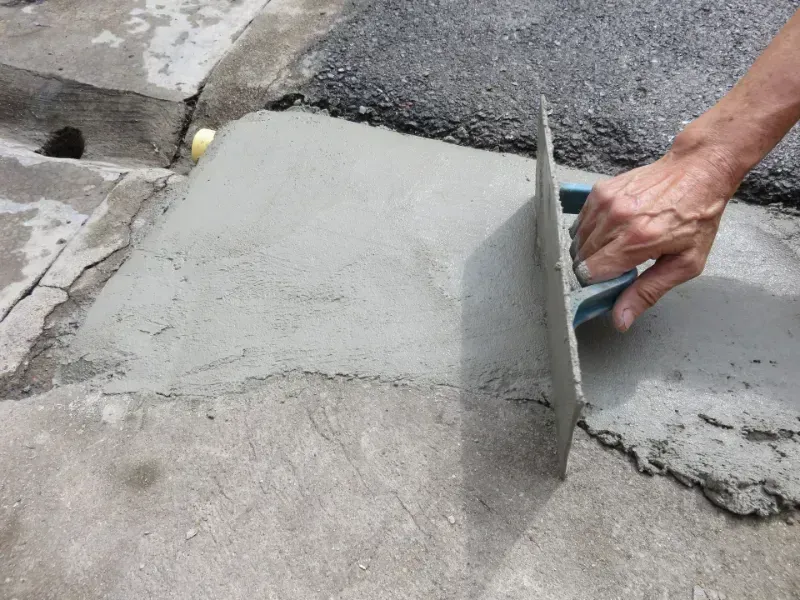 Worker smoothing fresh concrete with a hand trowel while repairing a sidewalk curb edge.