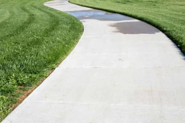 A paved concrete pathway curves through a grassy park area with a small puddle on the path.