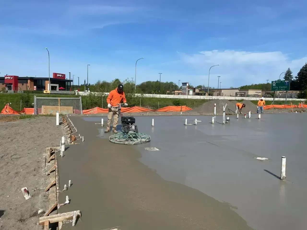 A construction worker uses a power trowel to smooth a large wet concrete slab on an outdoor job site under a clear sky.