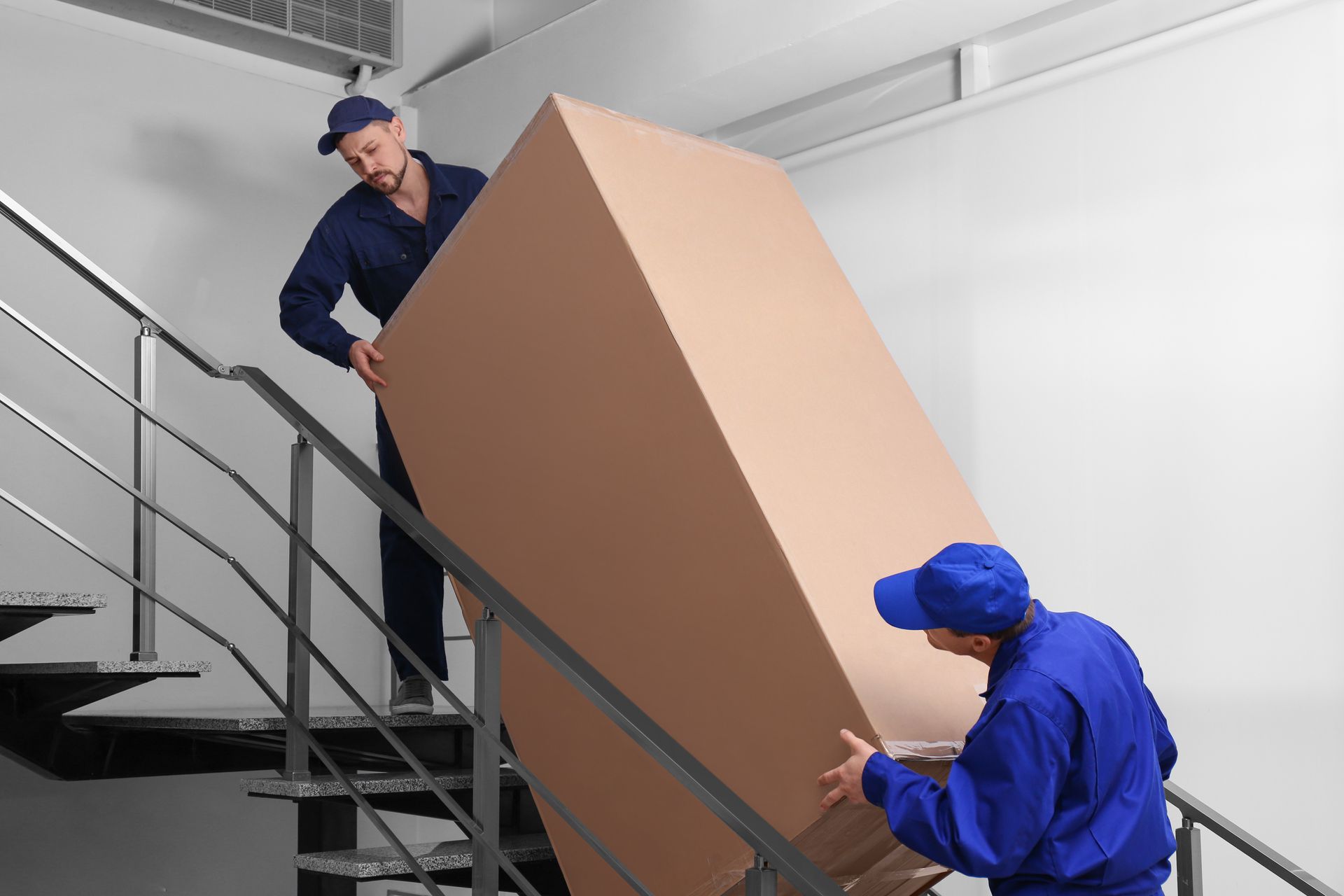 Two men are carrying a large cardboard box up a set of stairs.