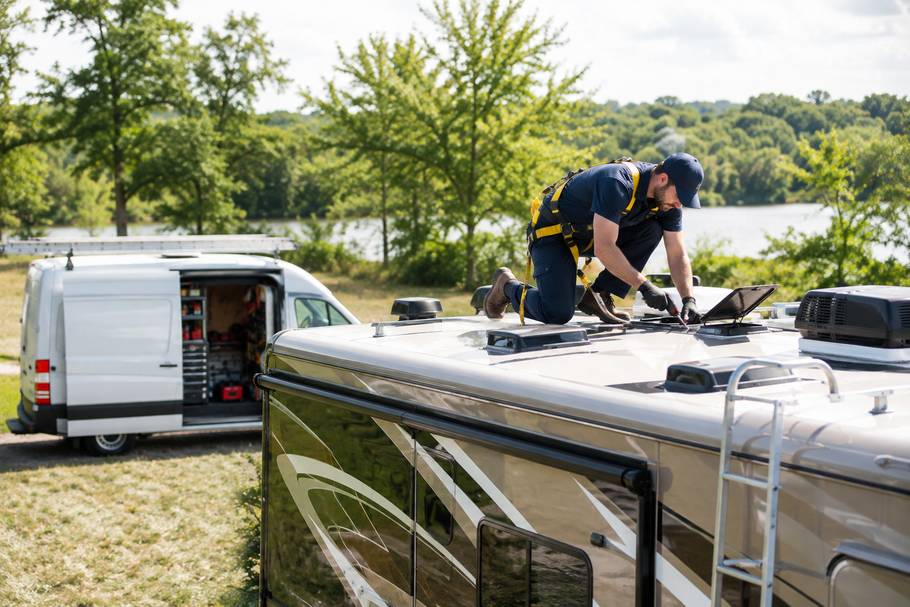 Worker on a van roof repairing equipment beside a white service van near water.