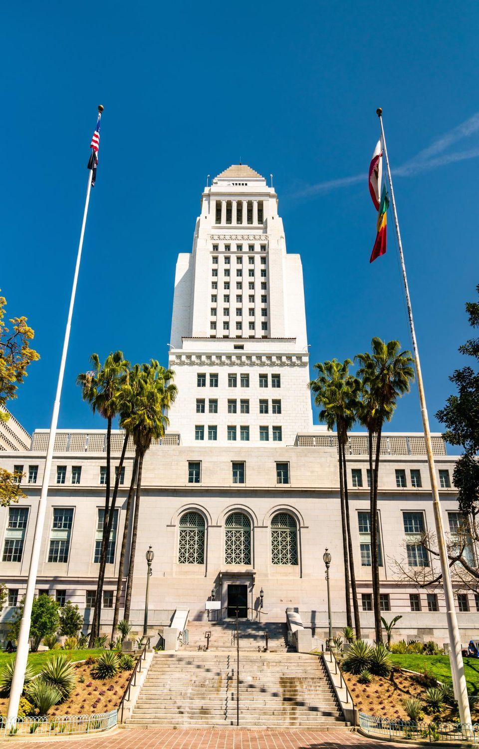 Tall white civic building with flagpoles and palm trees under a clear blue sky
