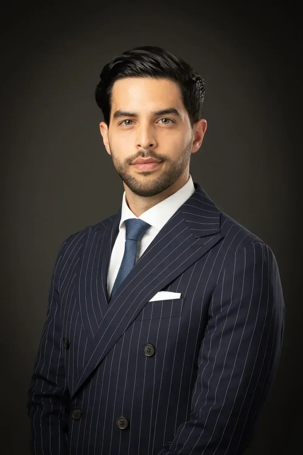 Studio portrait of a man in a navy pinstripe suit with a tie and pocket square on a dark background