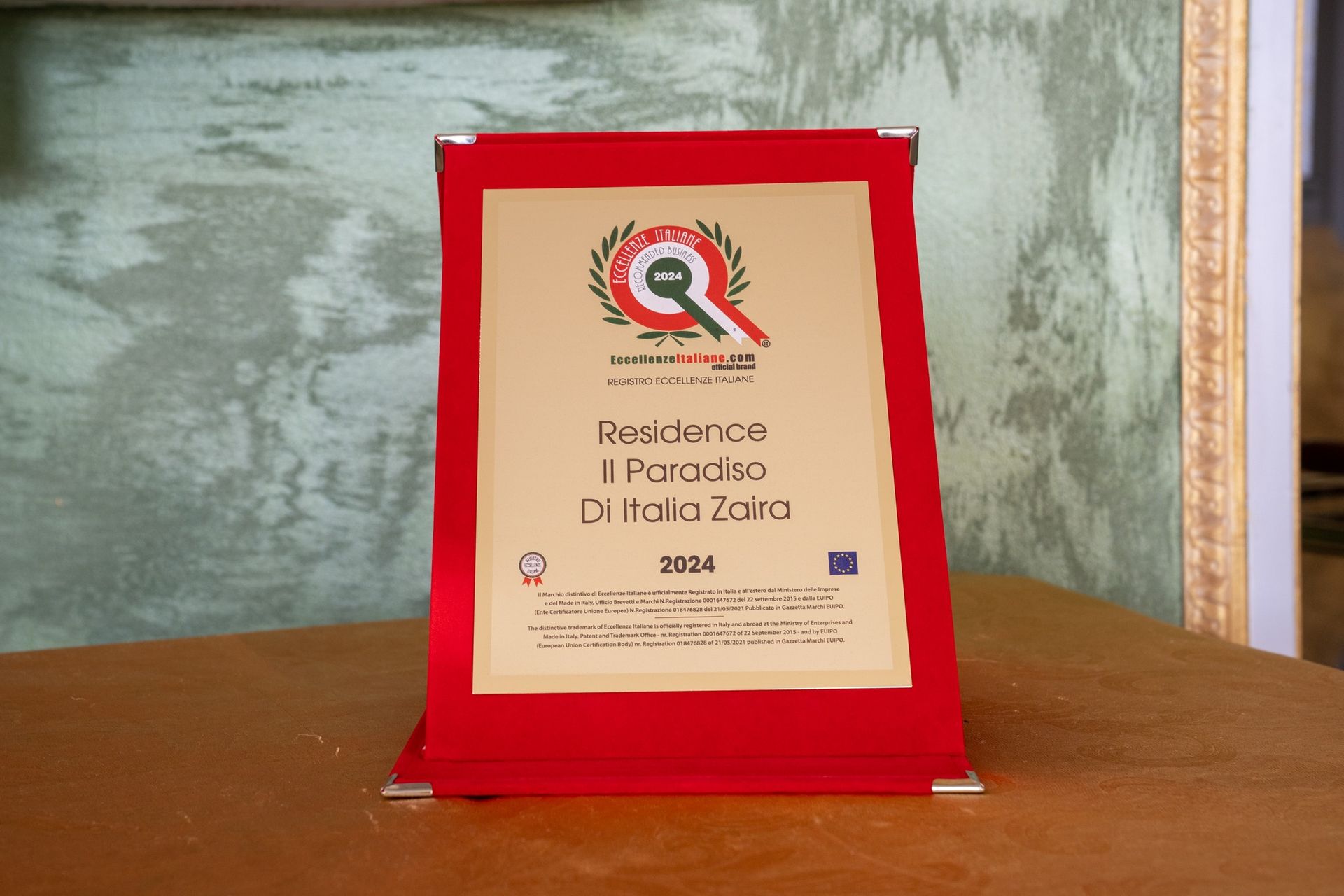 Un certificato incorniciato su un tavolo di legno. È rosso, con testo e logo in oro e bianco. Il certificato è per