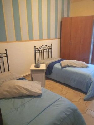 Due letti singoli con lenzuola blu in una stanza con carta da parati a righe blu e bianche, un comodino e un armadio marrone.