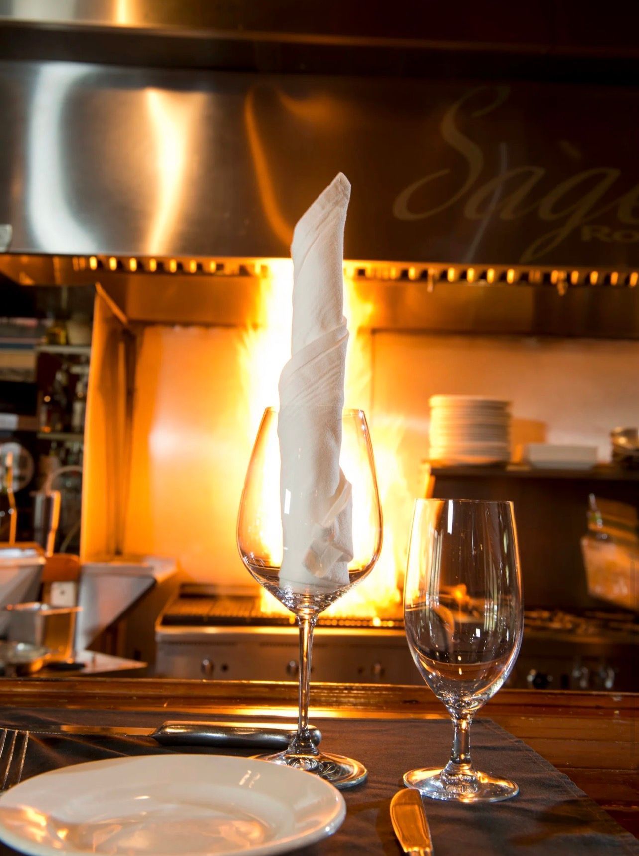 Place setting, including wine glass with napkin and grill in background.