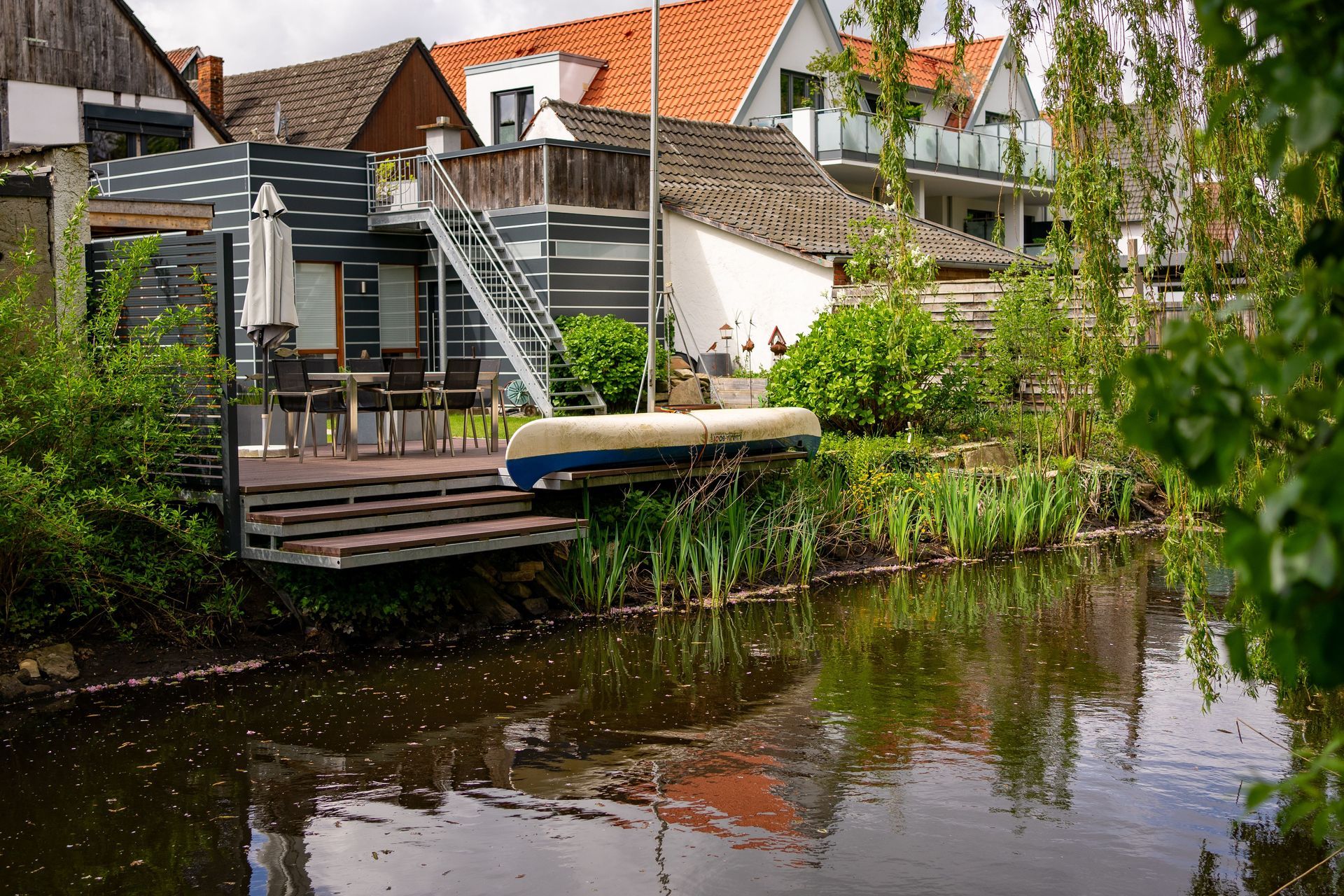 Canal-side house deck with white chairs, stairs, and greenery reflected in calm water