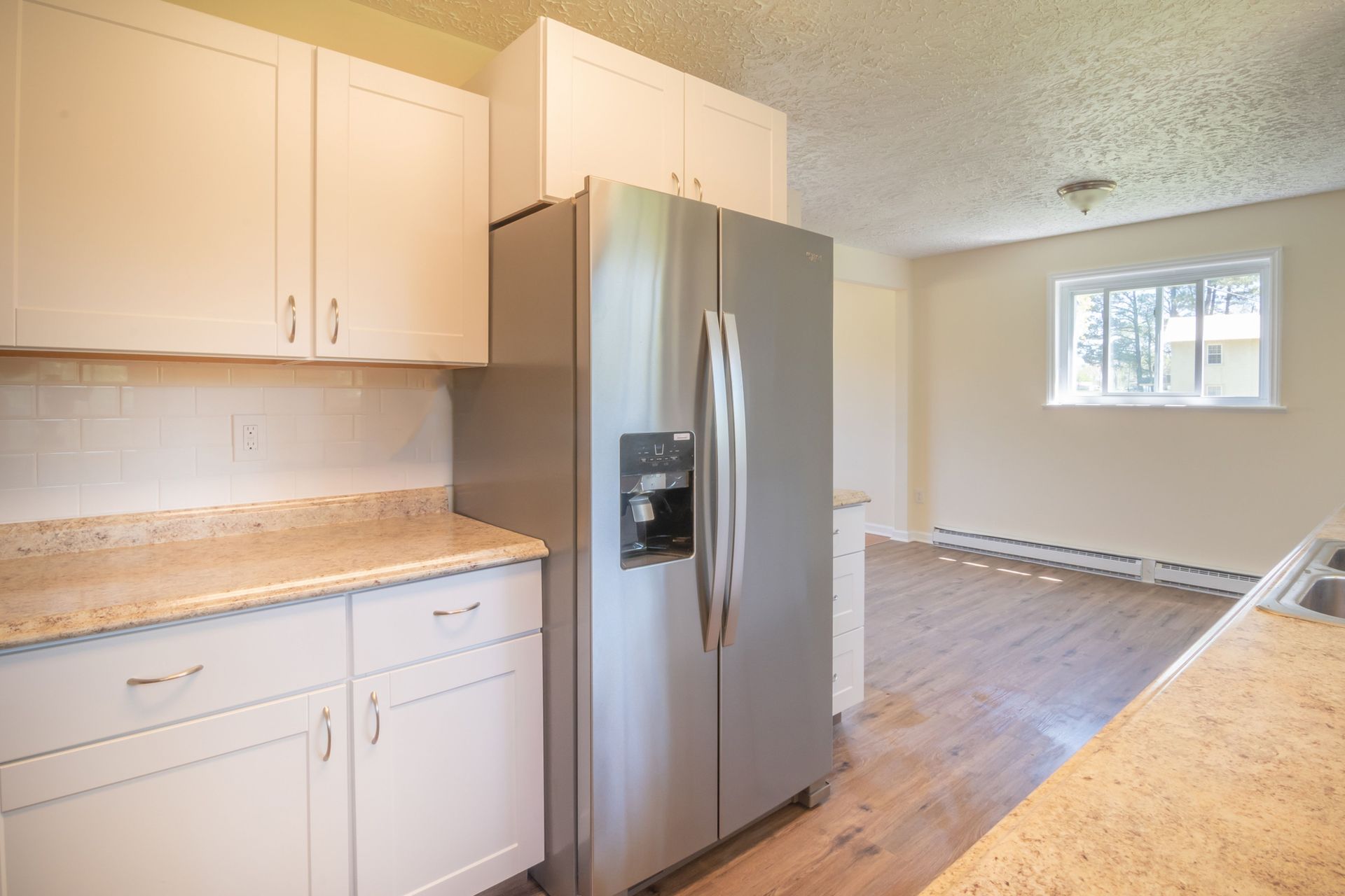 Bright kitchen with white cabinets, stainless steel fridge, and light wood flooring, opening into a sunlit room