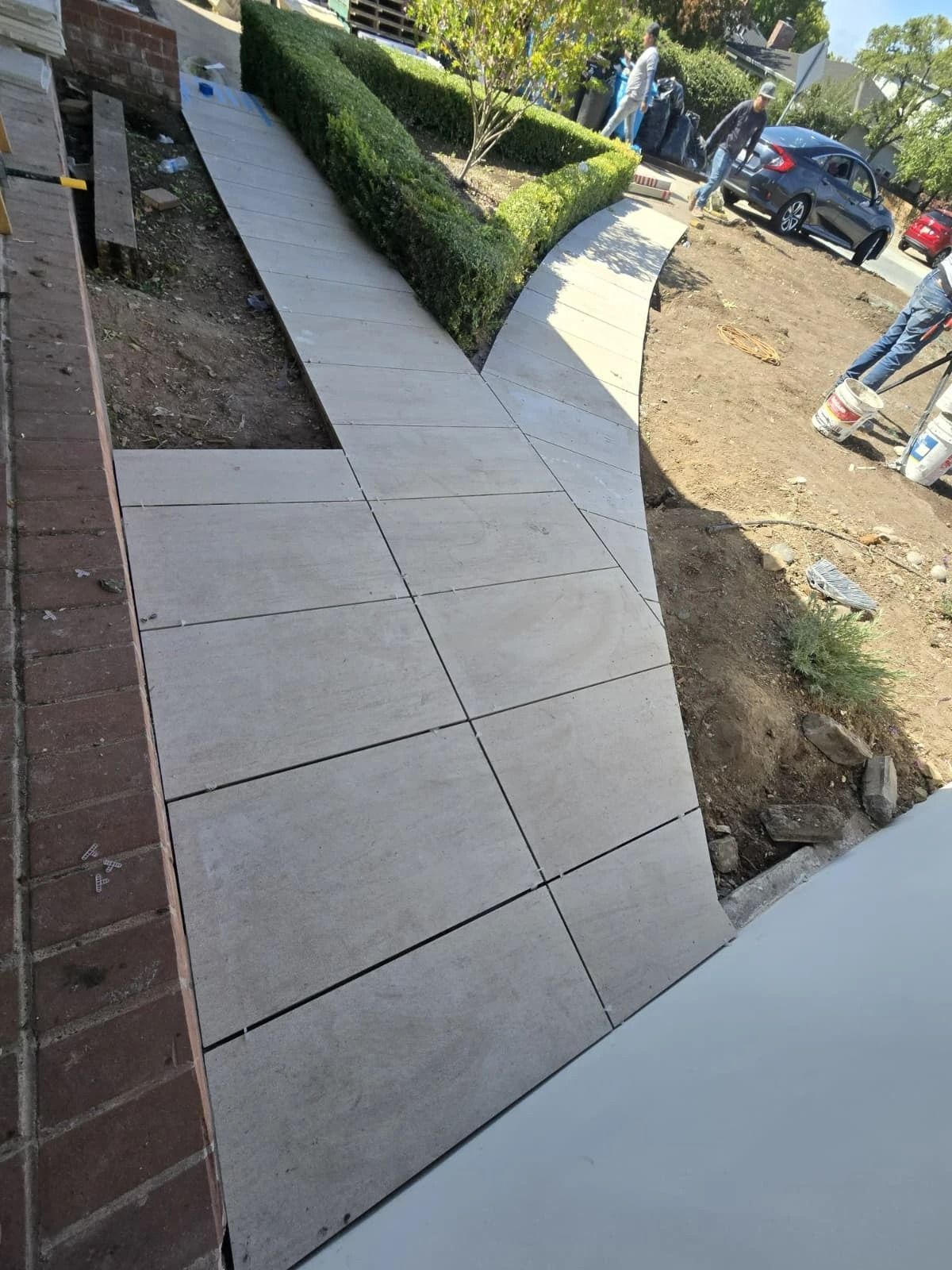 Sidewalk with newly laid concrete slabs beside a brick wall and trimmed hedges, near a street and parked car.