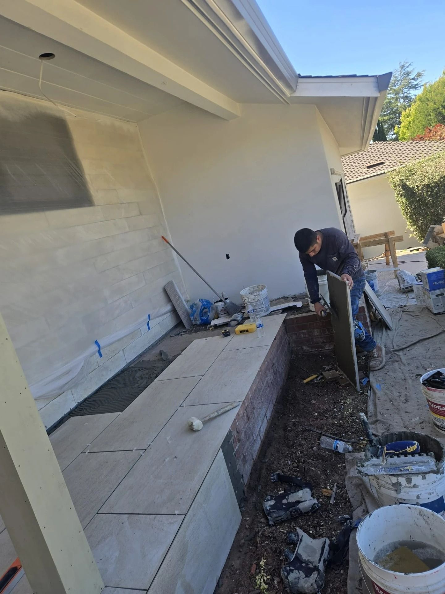 Worker repairing an outdoor patio beside a white house, with tools and construction materials nearby