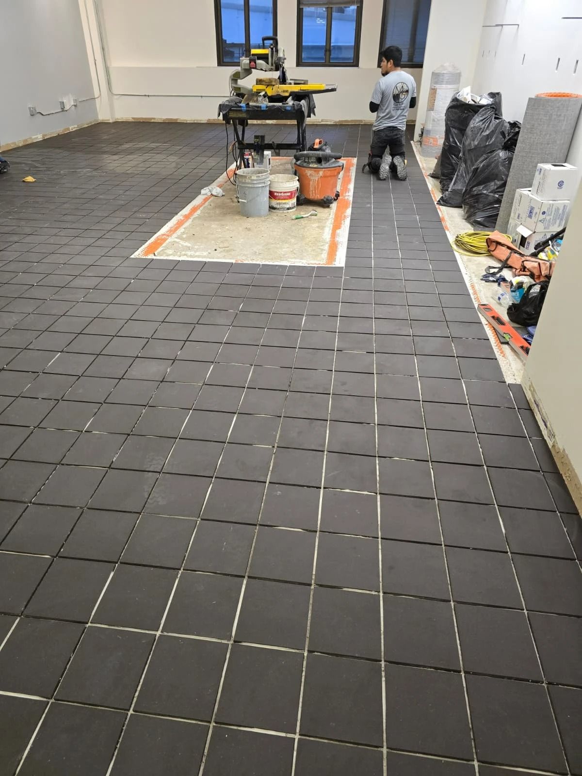 Large room under renovation with dark floor tiles, tools, and a worker near the back wall.