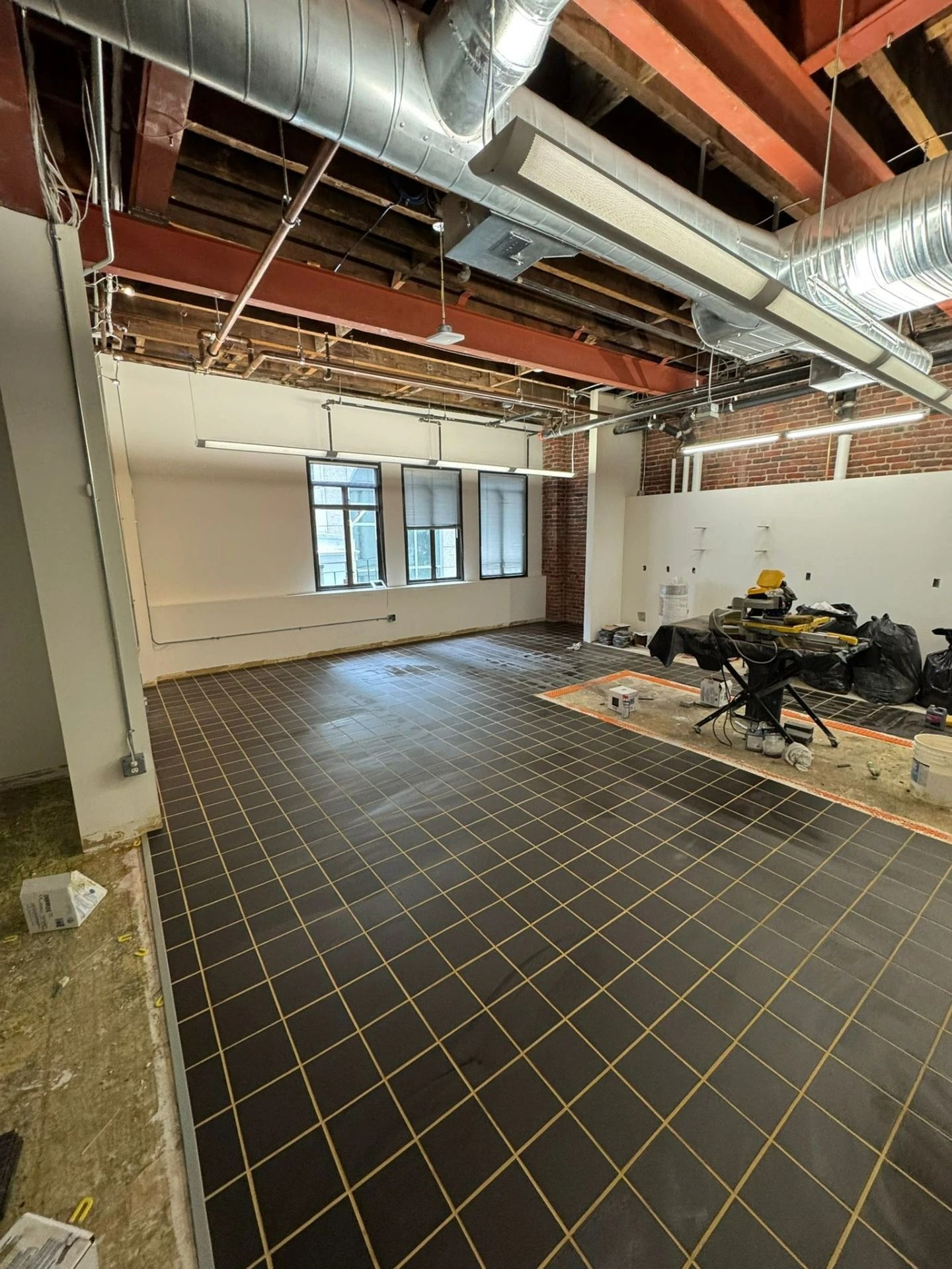 Empty industrial room with exposed ceiling beams, black tiled floor, and windows along one wall