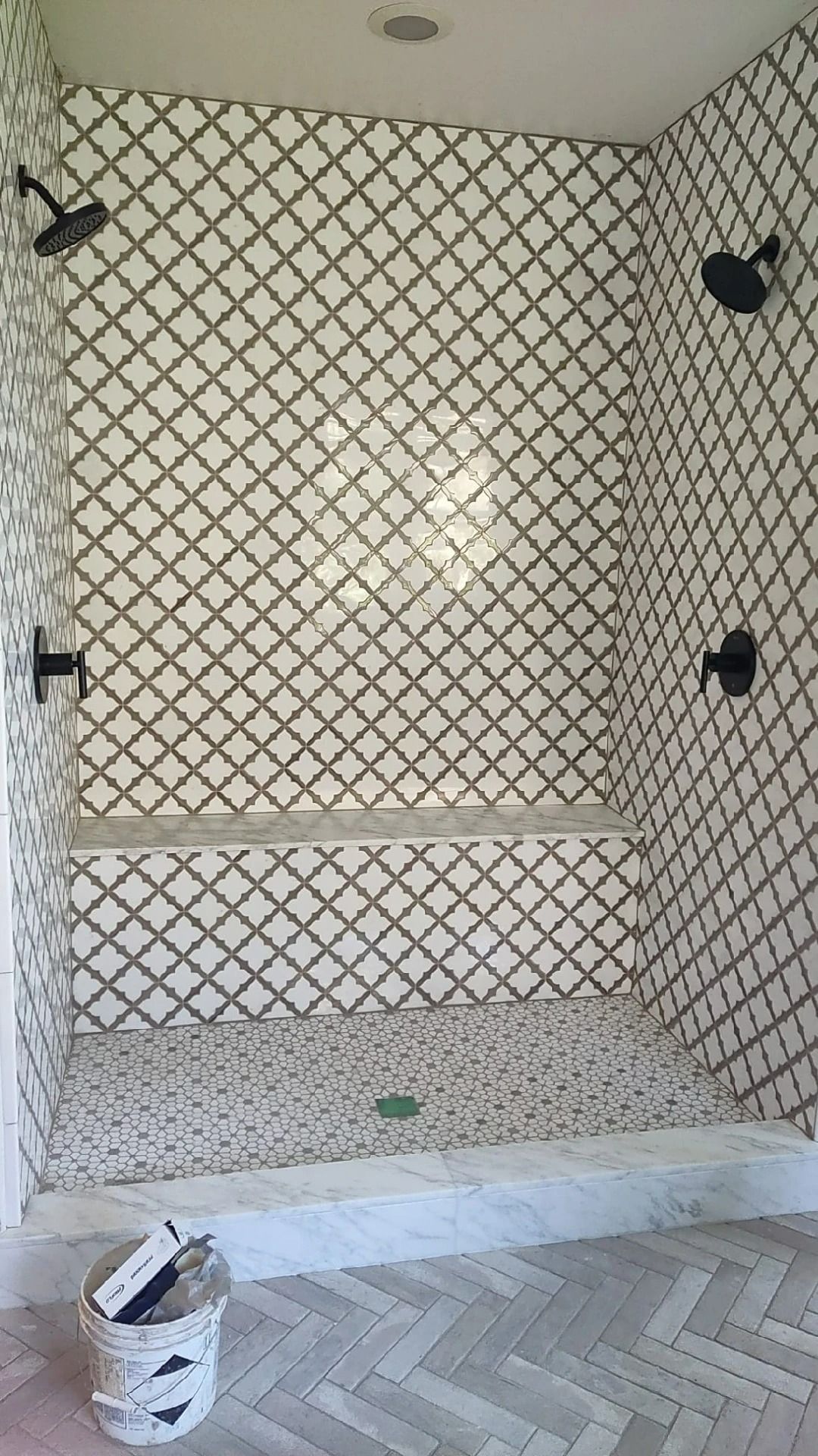 Tiled shower stall with black corner fixtures and a small white bucket on the floor