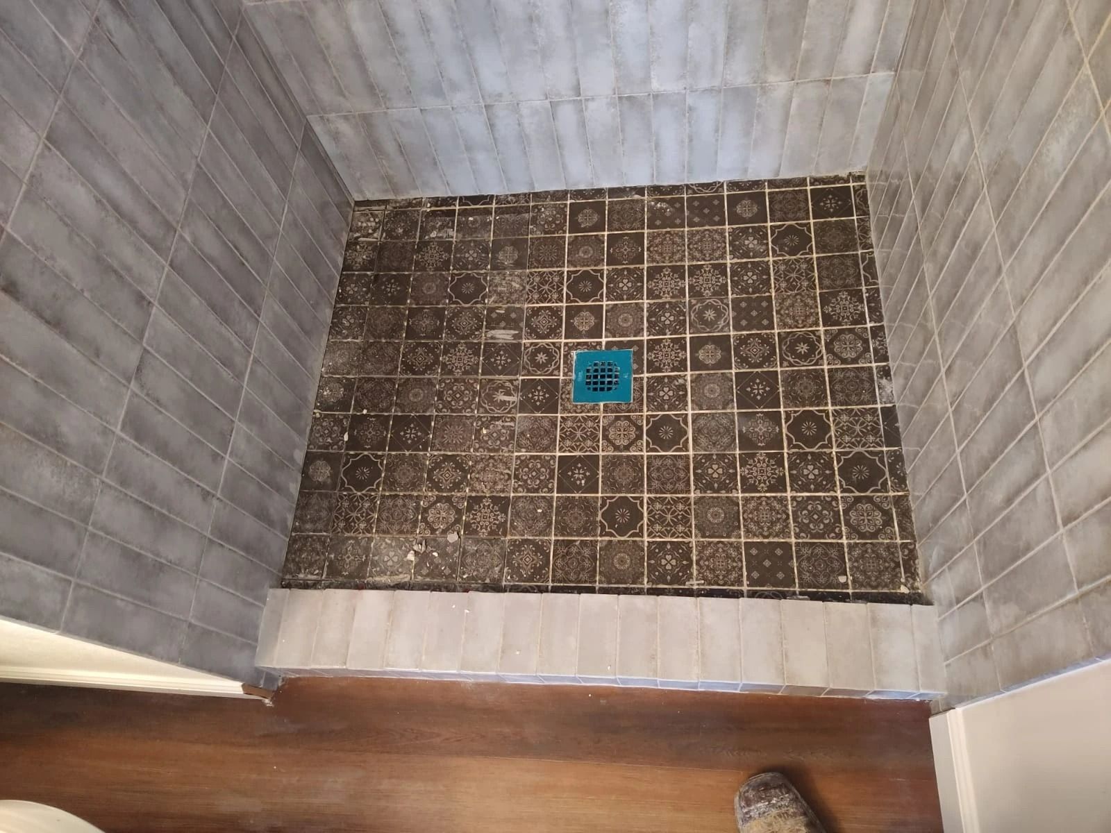 Empty tiled shower stall with a blue drain cover and wood floor visible at the entrance